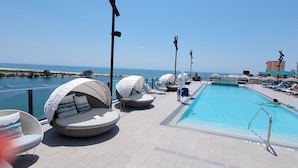 A poolside area with sun loungers and umbrellas, a clear view of the sea, and a tiled deck.