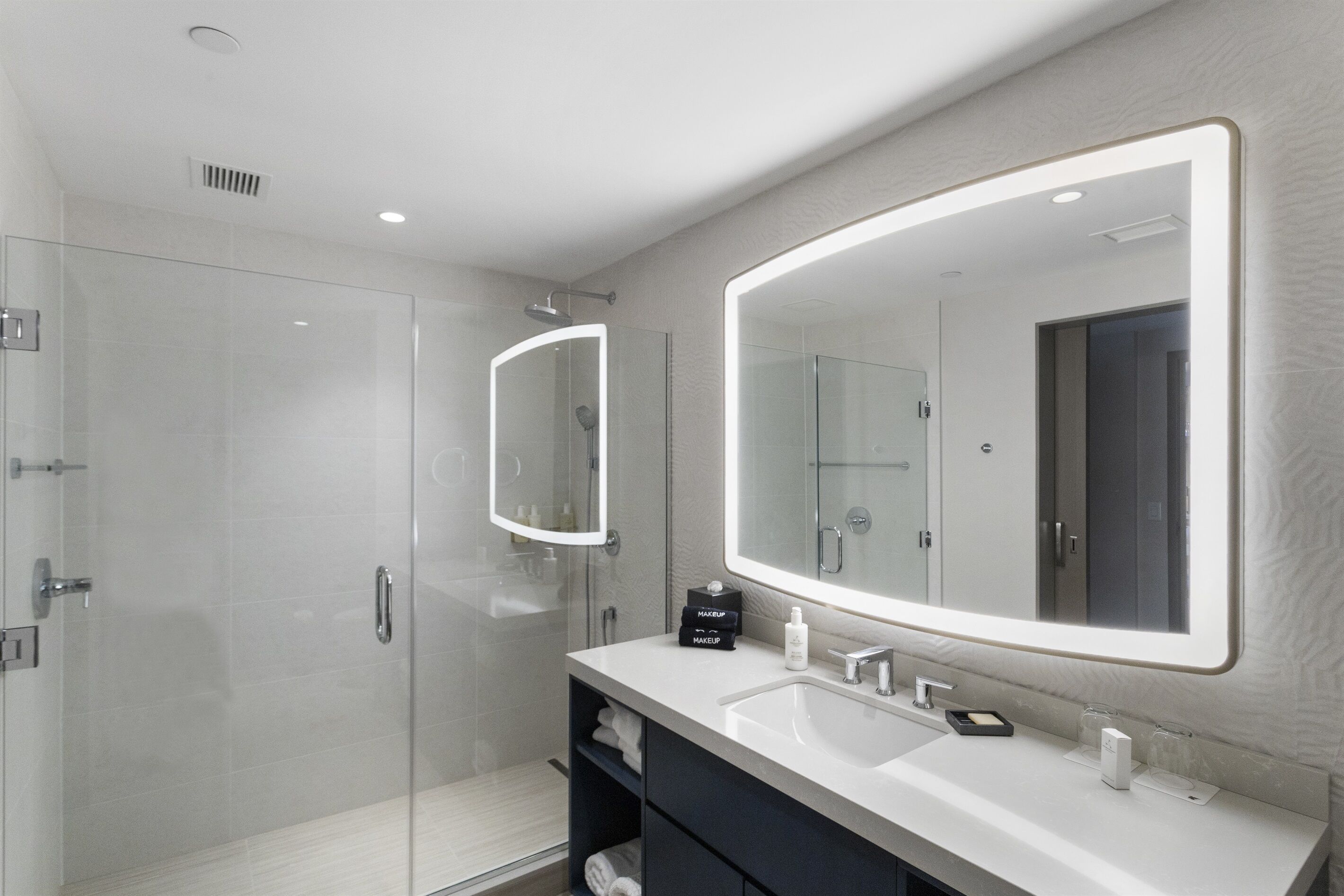 Un baño moderno con ducha envuelta en vidrio, un espejo grande y un lavamanos con almacenamiento.