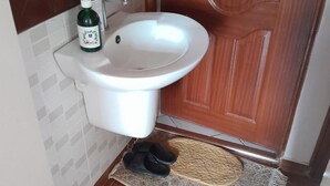 Une salle de bain avec un lavabo blanc, un flacon de savon liquide vert et une paire de pantoufles noires posées sur un tapis tissé.