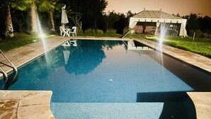 En swimmingpool med flisebelagt areal, et overdækket opholdsområde og en pavillon i baggrunden.