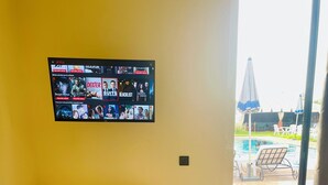Et hotelværelse med et fjernsyn, der viser forskellige programmer, et hvidt bord med en fjernbetjening, et lille rundt bord med en fjernbetjening og et rødt håndklæde på sengen.