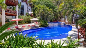 A swimming pool with lounge chairs and umbrellas, surrounded by lush greenery and a multi-story building with balconies. - The Sweet Life Sayulita | Ocean Views (Sayulita)