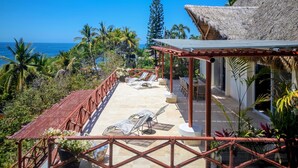 A covered outdoor seating area with chairs and tables, overlooking a beach with palm trees and a clear blue sky. - The Sweet Life Sayulita | Ocean Views (Sayulita)