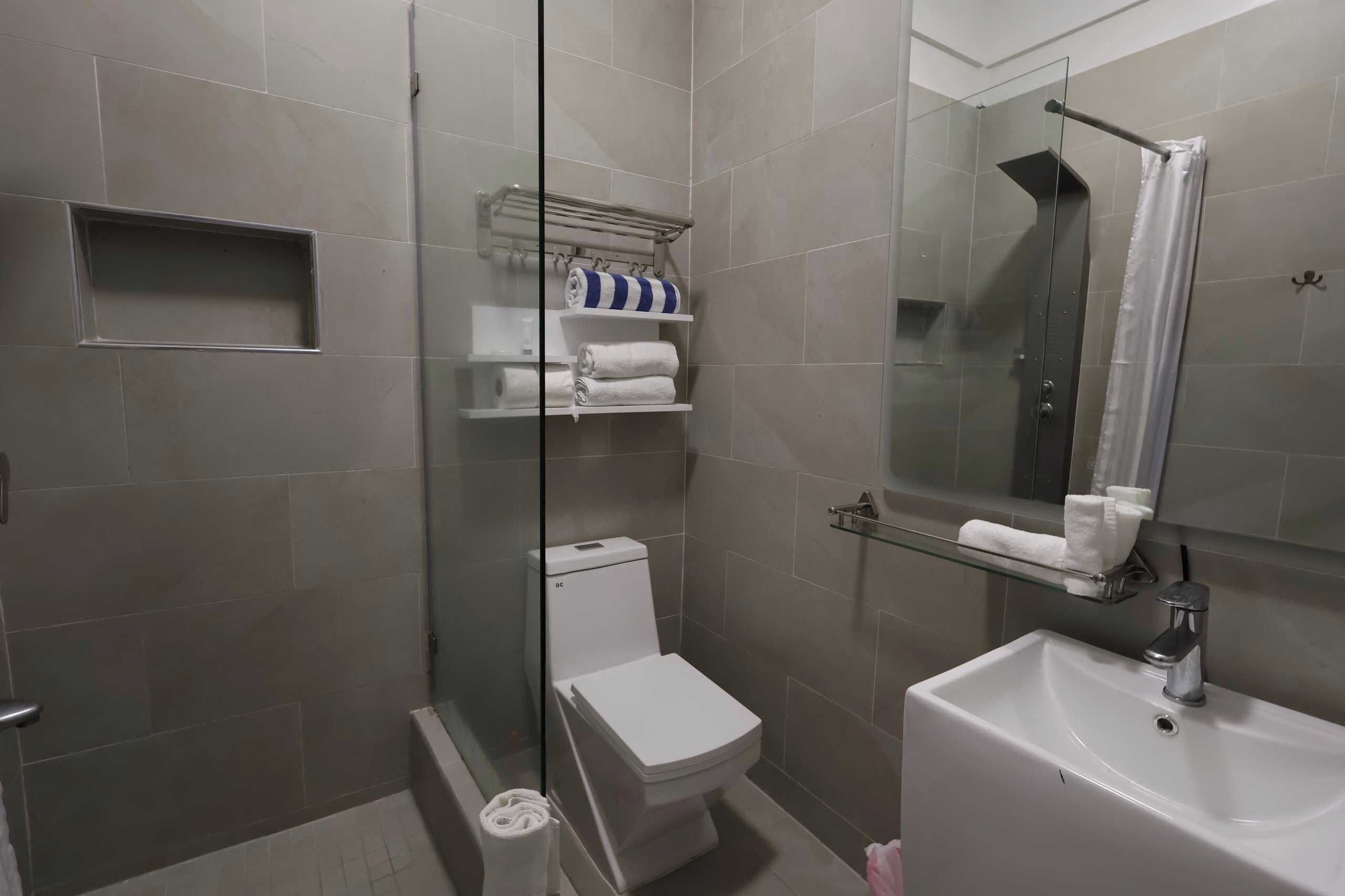 Un baño con ducha de vidrio, inodoro blanco, lavabo con espejo y repisas con toallas.