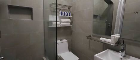 Un baño con ducha de vidrio, inodoro blanco, lavabo con espejo y repisas con toallas.