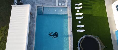 Vista aérea de una piscina con el logo de un tiburón, un jacuzzi y un área verde con sillas de descanso.