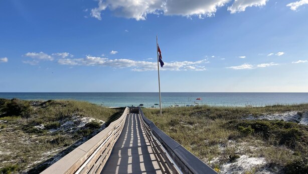 Une promenade en bois mène à une plage, et on aperçoit un mât de drapeau au loin.