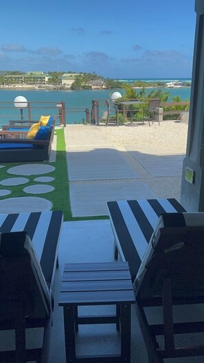 Ein Strandloungebereich mit Liegestühlen und einem kleinen Tisch, mit Blick auf ein klares blaues Meer und eine entfernte Küste mit Gebäuden.