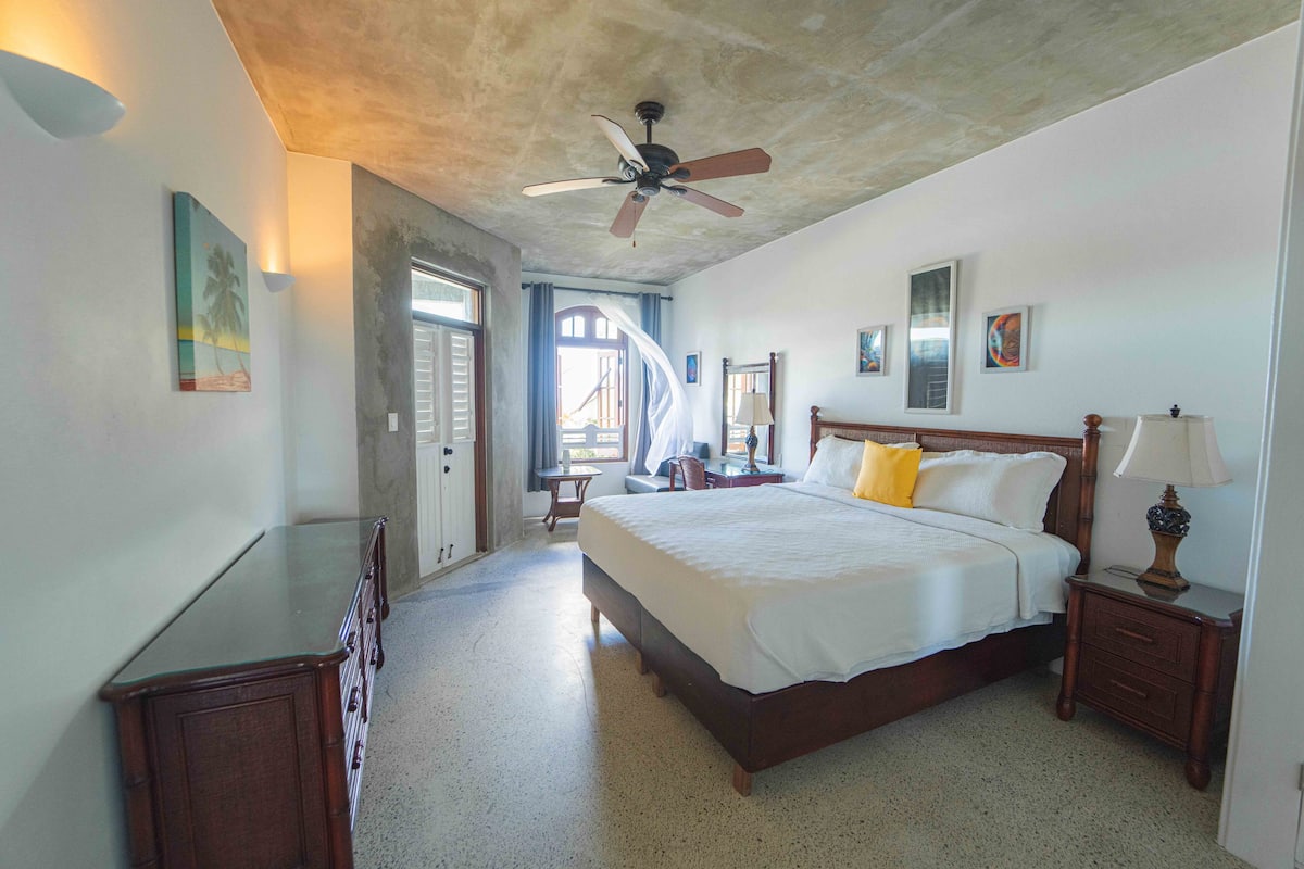 A spacious bedroom with a large bed, a ceiling fan, and a view of the outside.