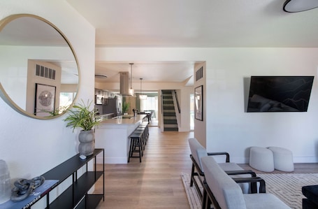 A modern living room with a central island, a staircase, and a kitchen in the background.