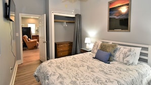 A bedroom with a bed, a nightstand, a lamp, a painting, a ceiling fan, a wardrobe, and a chair. - Teal Door Inn-With Fire Pit! (Charleston)