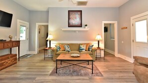 A living room with a sofa set, two side tables with lamps, a coffee table, a ceiling fan, a mounted TV, and a framed picture on the wall. - Teal Door Inn-With Fire Pit! (Charleston)