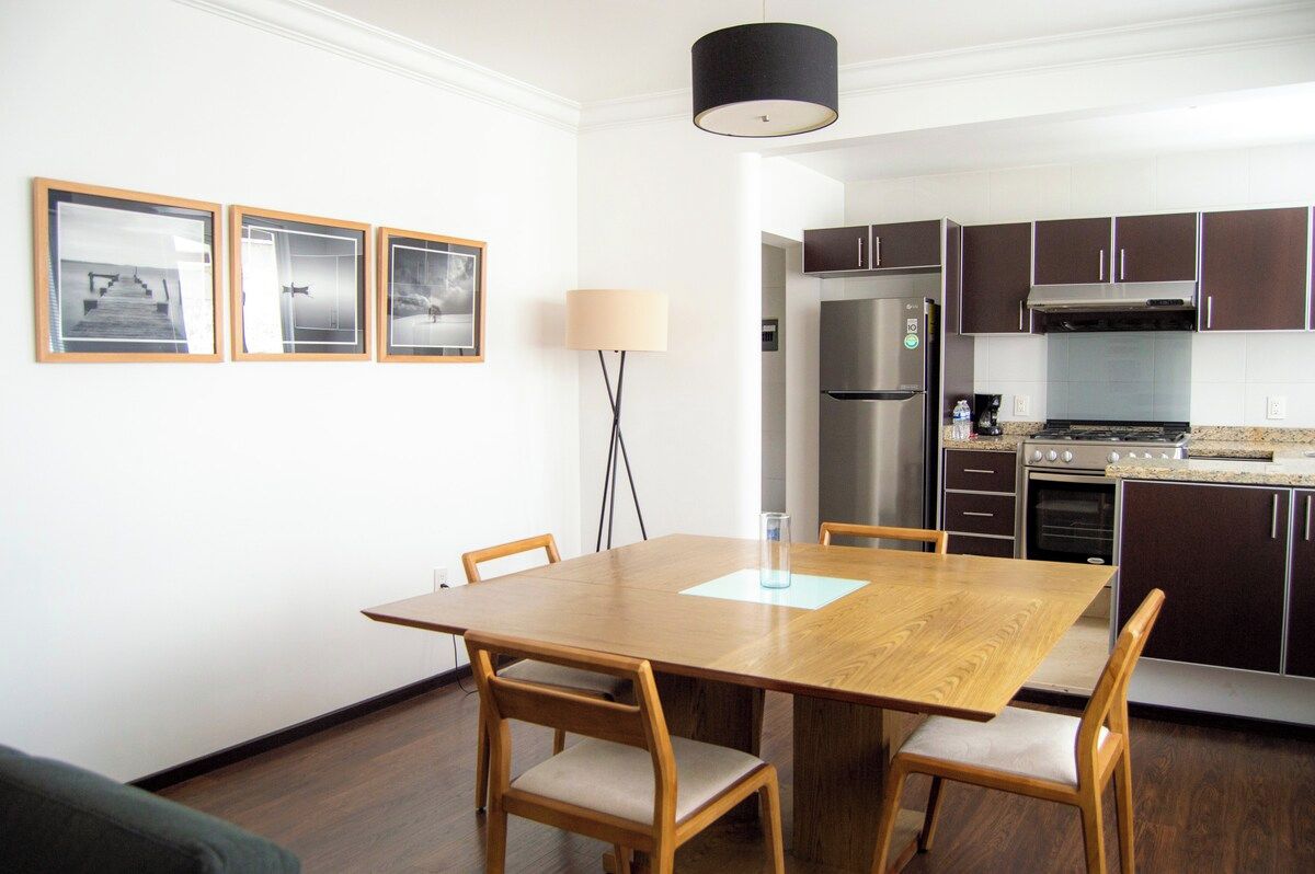 Une cuisine moderne avec des meubles de couleur sombre, des appareils en acier inoxydable et une table à manger en bois, accompagnée de chaises.