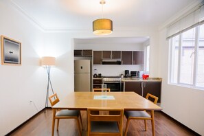 Zona pranzo con tavolo e sedie in legno, una lampada a sospensione e una cucina sullo sfondo.