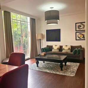 Ein Wohnzimmer mit einer Couch, einem Couchtisch und einer Lampe. Ein Fenster mit Vorhängen, ein Gemälde an der Wand und ein gemustertes Teppich sind ebenfalls vorhanden.