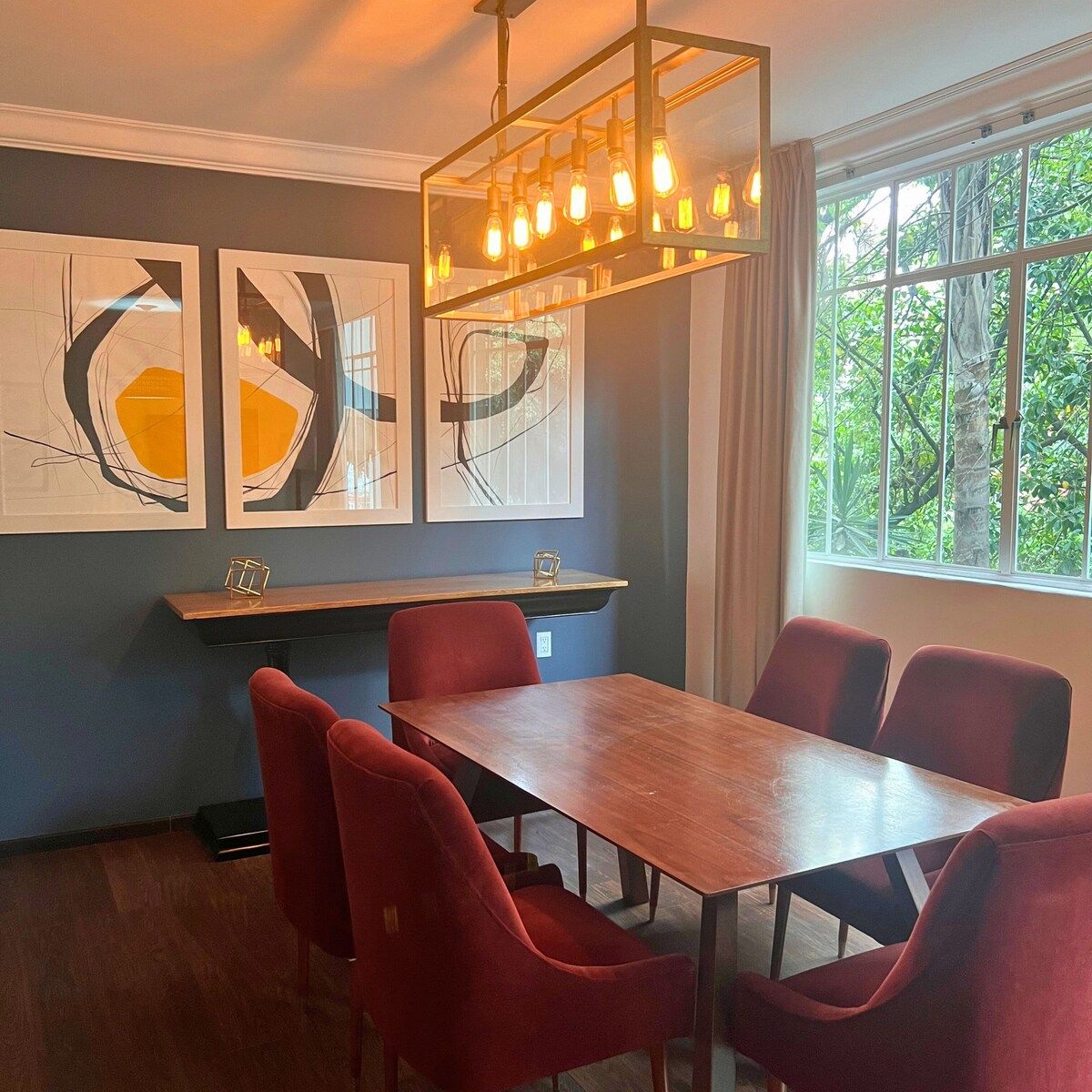 Un coin repas avec une table en bois et des chaises rouges, des œuvres d’art encadrées accrochées au mur et une fenêtre donnant sur un paysage verdoyant.