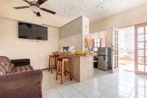 A living room with a sofa, a TV mounted on the wall, a kitchen area with a refrigerator, and a ceiling fan. - Shaw Park Gem Apt 12A (Ocho Rios)