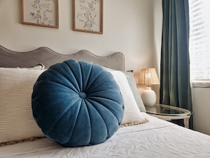 Un lit bien fait, agrémenté d’un grand oreiller bleu, d’une couette blanche et d’une tête de lit. Deux reproductions botaniques encadrées ornent le mur, une lampe se trouve sur une table de chevet, et une fenêtre est habillée de rideaux.