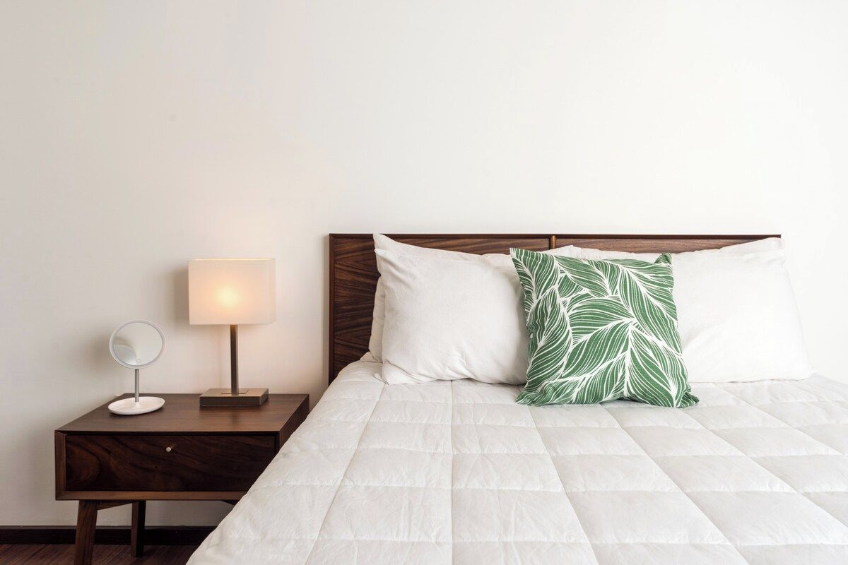 A neatly made bed with a white duvet, a wooden headboard, and a green leaf-patterned pillow.