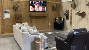 A spacious room with a large flat-screen TV mounted on a wooden wall, a grey sectional sofa, a black leather recliner, and several mounted animal heads.