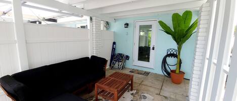 Un espacio de estar al aire libre techado, con un sofá negro, una mesa de madera, una planta en maceta y una puerta que da a un porche cubierto.