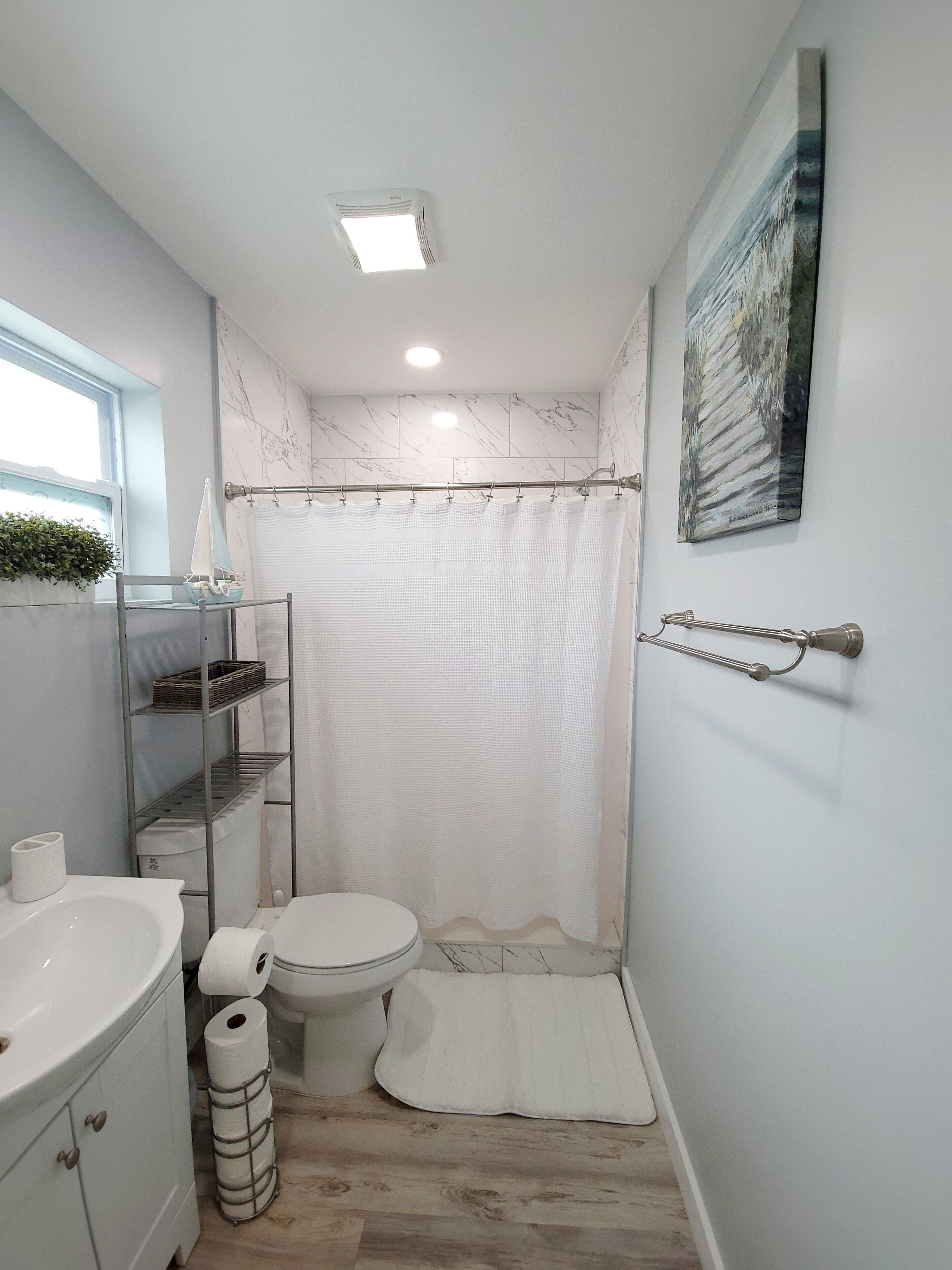 A compact bathroom with a toilet, a sink, a shower curtain, a shelf with toiletries, and a window with a plant.