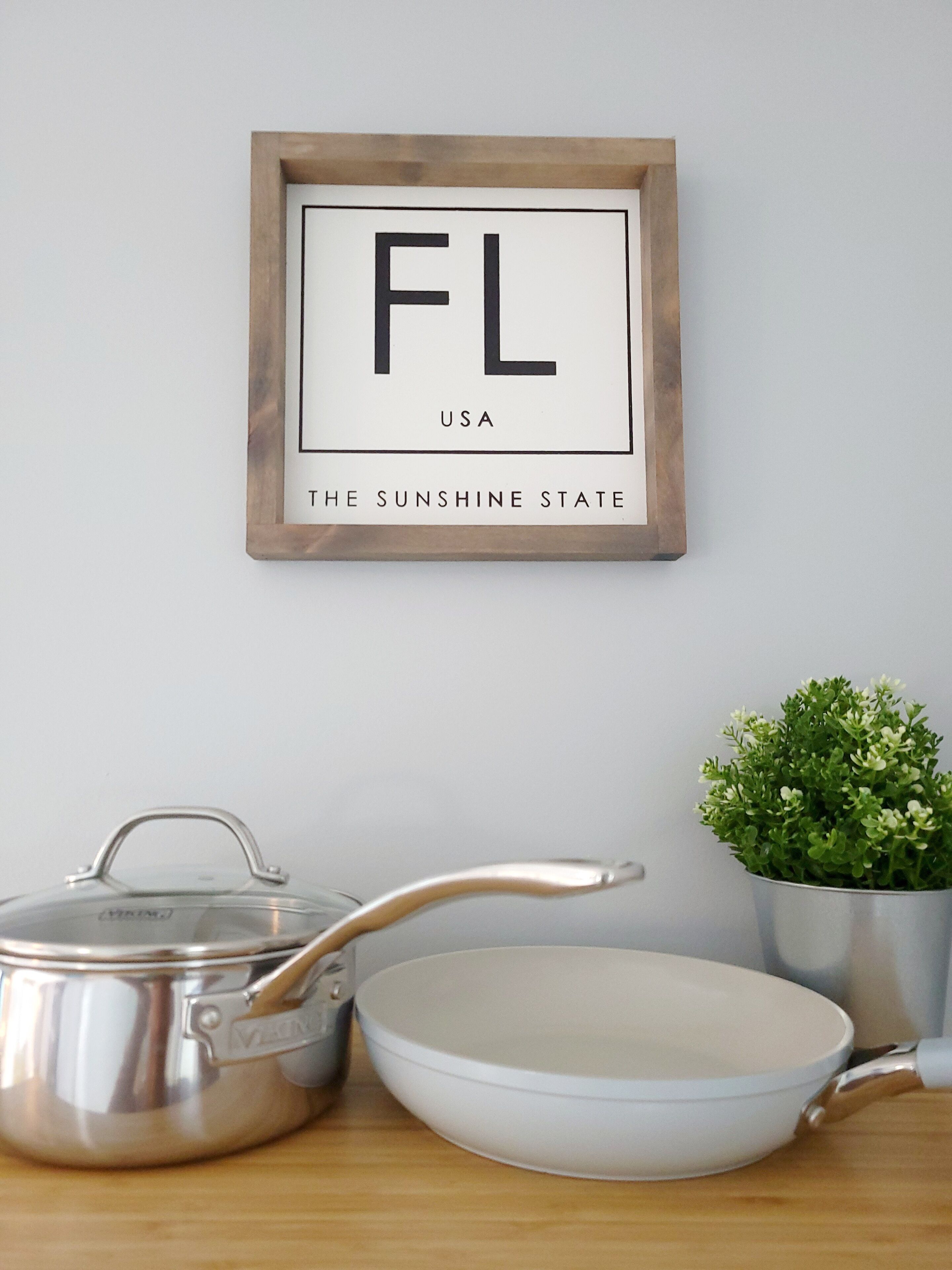 A framed sign with "FL" and "USA" on a wall, a metal pot, a white bowl, and a small potted plant.
