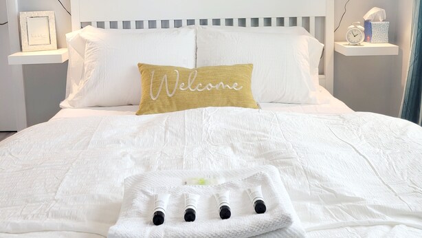 Un lit bien fait, avec une couette blanche, un coussin orné du mot «Bienvenue» et une pile d’essuie-serviettes.