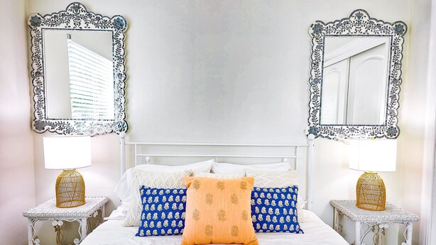 Ein ordentlich eingerichtetes Schlafzimmer mit einem weißen Bett, blau gemusterten Kissen und einem zentral platzierten orangefarbenen Kissen. Zwei verzierte Spiegel flankieren das Bett, und es gibt zwei kleine Nachttische mit Lampen.