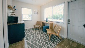 Eine kompakte Küchenzeile mit Mikrowelle, Kaffeemaschine und einer kleinen Ablage für Tassen. Ein Fenster mit Jalousien, eine Tür und ein gemustertes Teppich sind ebenfalls vorhanden.