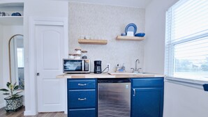 Eine kompakte Küche mit Mikrowelle, Kaffeemaschine und Geschirrspüler. Blaue Schränke und ein kleines Fenster mit Jalousien.