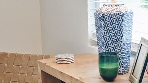 Ein Teil eines Raumes mit einem Holztisch, einem gewebten Stuhl, einer blauen gepolsterten Sitzbank, einem gemusterten Kissen und einem grünen Glas.