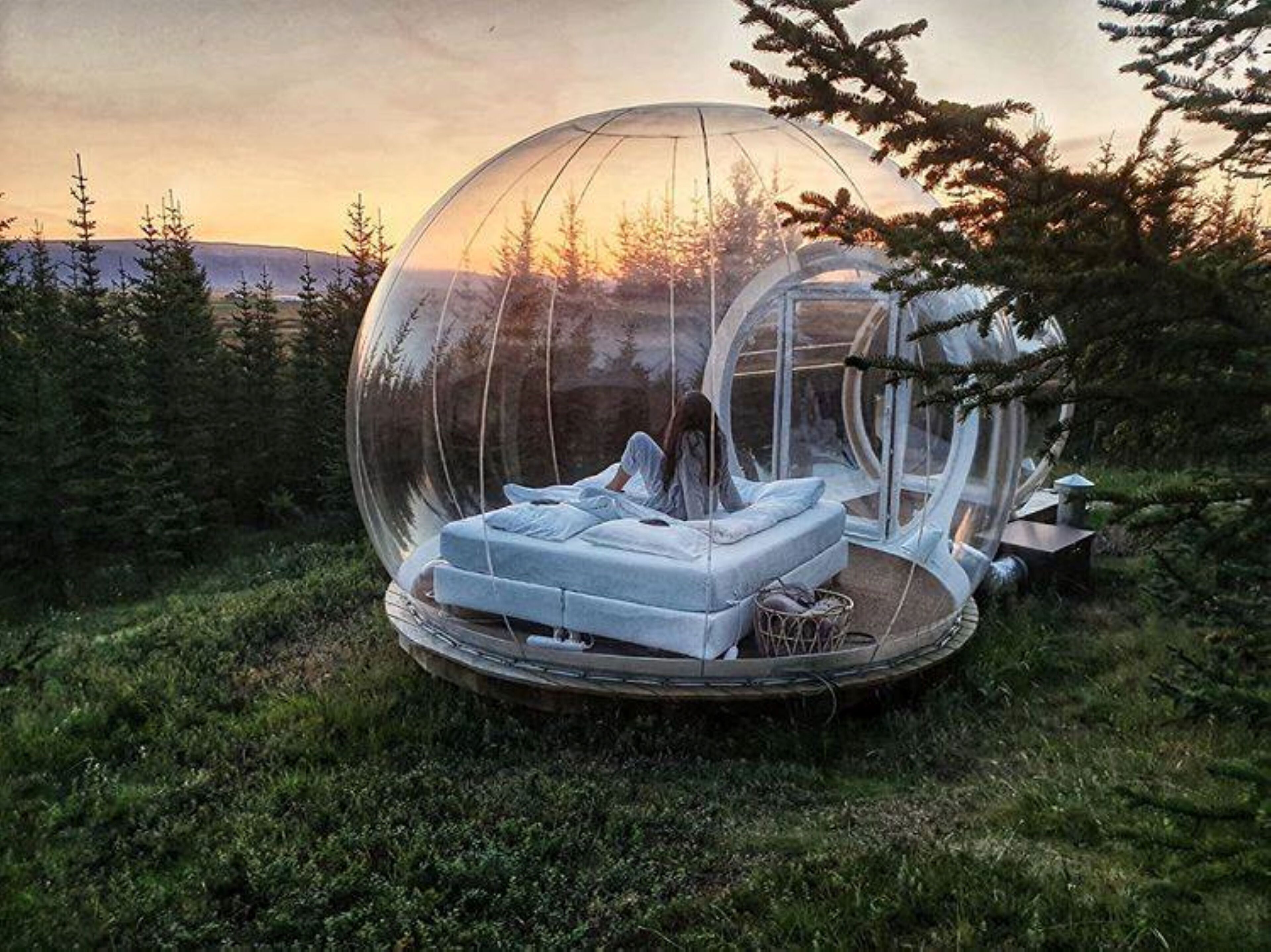 A transparent spherical structure with a bed inside, surrounded by trees and a sunset in the background.