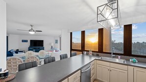 Een moderne keuken met een centraal eiland, een eethoek met een glazen tafel en uitzicht op de ondergaande zon door grote ramen.