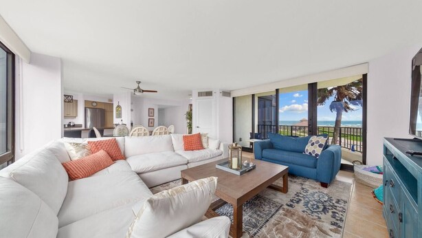 Een ruime woonkamer met een witte hoekbank, een blauwe bank en een houten salontafel. Er is een balkon met uitzicht op de oceaan.