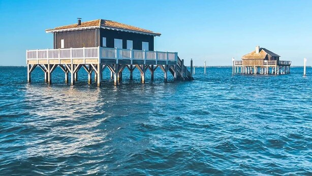 Zwei hölzerne Konstruktionen auf Stelzen über ruhigem Wasser, vermutlich als Unterkunft oder Speisegaststätte genutzt.