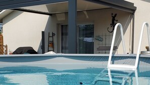 Ein Swimmingpool mit weißer Leiter, eine überdachte Terrasse und ein Gebäude mit weißer Fassade und braunem Dach.