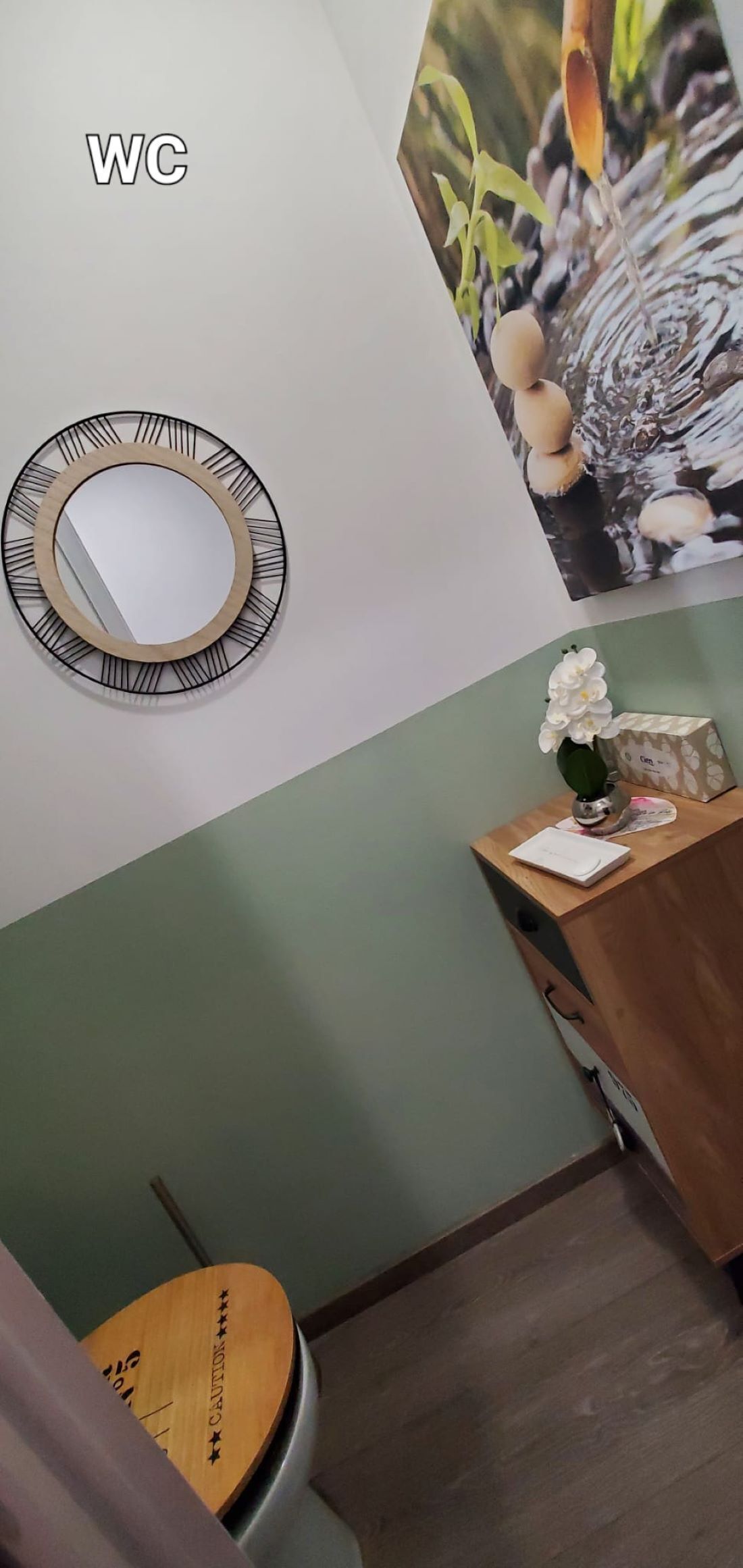 Une salle de bain avec des toilettes en bois, un miroir avec un cadre noir, un lavabo fixé au mur et un panneau d’avertissement sur le couvercle des toilettes.
