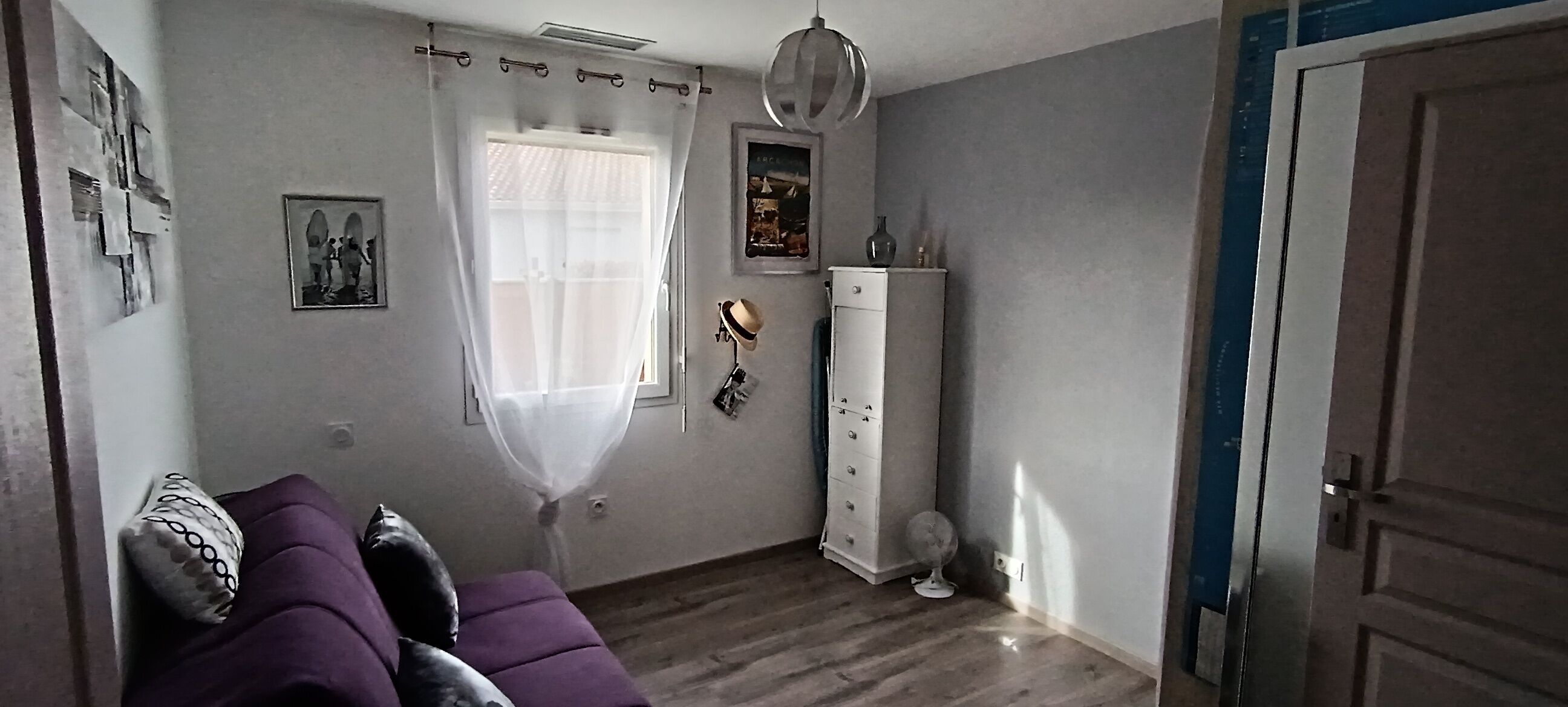Een kamer met een paarse bank, een wit kastje, een ventilator en een deur.