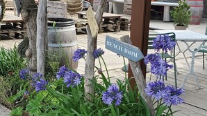 Ein hölzernes Wegweiser mit Richtungsschildern, ein Holzfass und violette Blumen in einem Garten.
