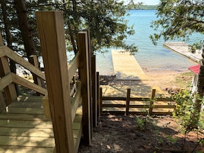 Eine Holzterrasse führt zu einem Strand, wo ein Steg in einen See hineinreicht, umgeben von Bäumen.