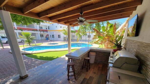 Un espace extérieur abrité comprenant une piscine, un sol en bois, un bar avec des tabourets et un barbecue.
