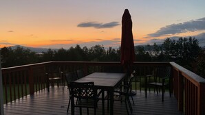 Eine Holzdielen-Terrasse mit Tisch, Stühlen und Sonnenschirm, mit Blick auf den Sonnenuntergang über einer bewaldeten Landschaft.