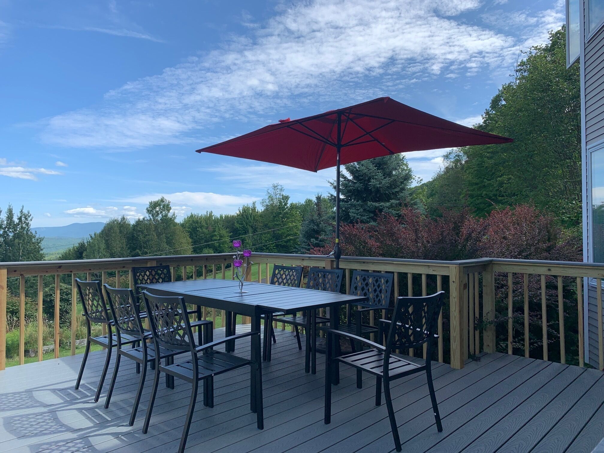 A deck with a table and chairs, a red umbrella, and a view of trees and mountains.