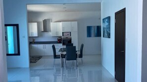 Une cuisine moderne avec des meubles blancs, un coin repas doté d’une table et de chaises en verre, et une porte noire.