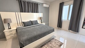 A modern bedroom with a large bed, bedside tables, a nightstand, a window with curtains, and an air conditioning unit. - PÀRIS Villas 3BD Villa Rodney Bay (Gros Islet)