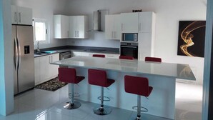 Une cuisine moderne avec des armoires blanches, un réfrigérateur en acier inoxydable, un four à micro-ondes et une cuisinière à gaz. On trouve trois tabourets de bar rouges autour d’un îlot central.