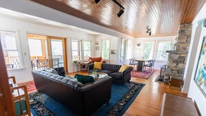 A spacious living room with a leather sofa, a coffee table, and a stone fireplace. There are multiple windows providing natural light and a view of the outdoors.