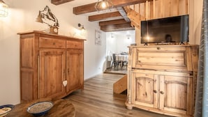 A wooden interior with a TV on a wooden cabinet, a clock, and a dining area in the background. - LA Petite Maison - Bonvillard (BONVILLARD)