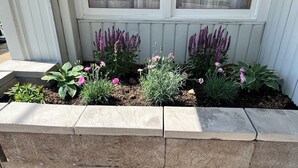 A raised garden bed with various plants and flowers, including purple flowers and green foliage, against a white wall with windows. - LakeWay II- 4 min from US-41 and Lake Sullivan (Sullivan)